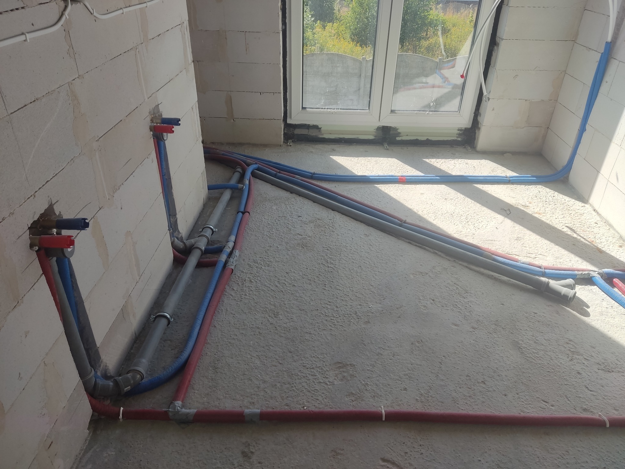 Wnętrze w trakcie prac instalacyjnych: widoczne rury instalacji wodnej w kolorach czerwonym i niebieskim, częściowo zamocowane do ściany z bloczków, oraz szare rury kanalizacyjne na surowym...