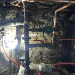 Nowe rury miedziane instalacji grzewczej w piwnicy z kamiennymi ścianami, z widocznym zaworem, pompą obiegową i elementami sterującymi na piecu.