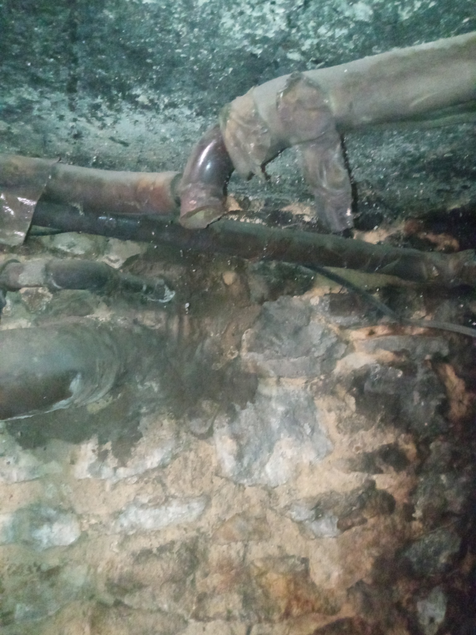 Stare, skorodowane rury hydrauliczne w ciemnym, wilgotnym otoczeniu, prawdopodobnie w piwnicy lub pod ziemią, z widocznymi śladami korozji i osadów na powierzchni.