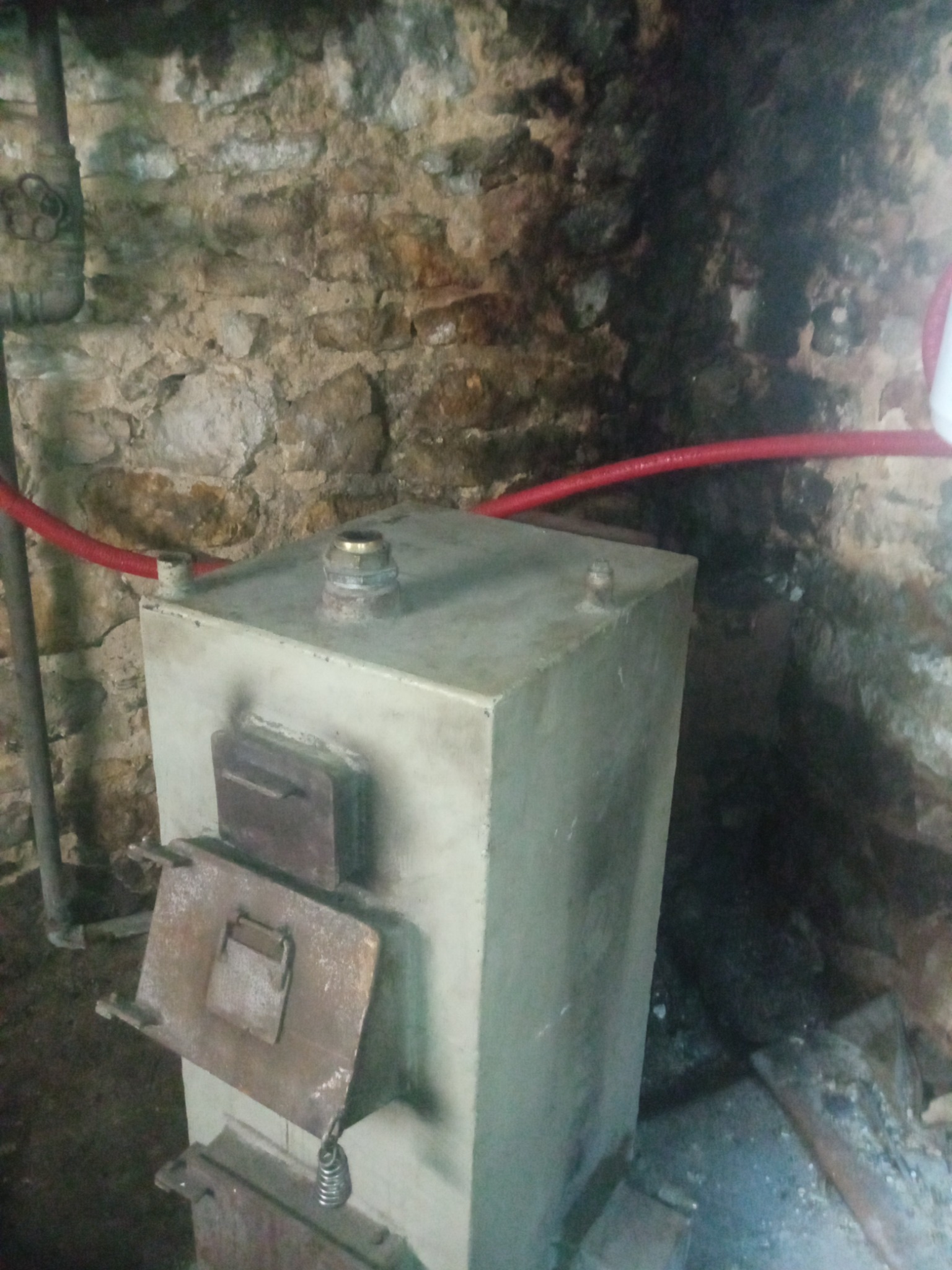 Metalowy piec z dwiema klapami w piwnicy o kamiennych ścianach, z widocznymi rurami instalacji grzewczej.