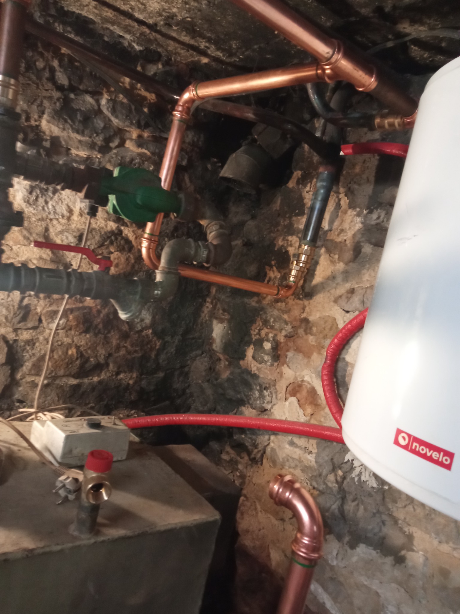 Nowe miedziane rury instalacji grzewczej i hydraulicznej na tle starej kamiennej ściany piwnicy, z widocznym bojlerem marki Novelo i elementami zaworów.