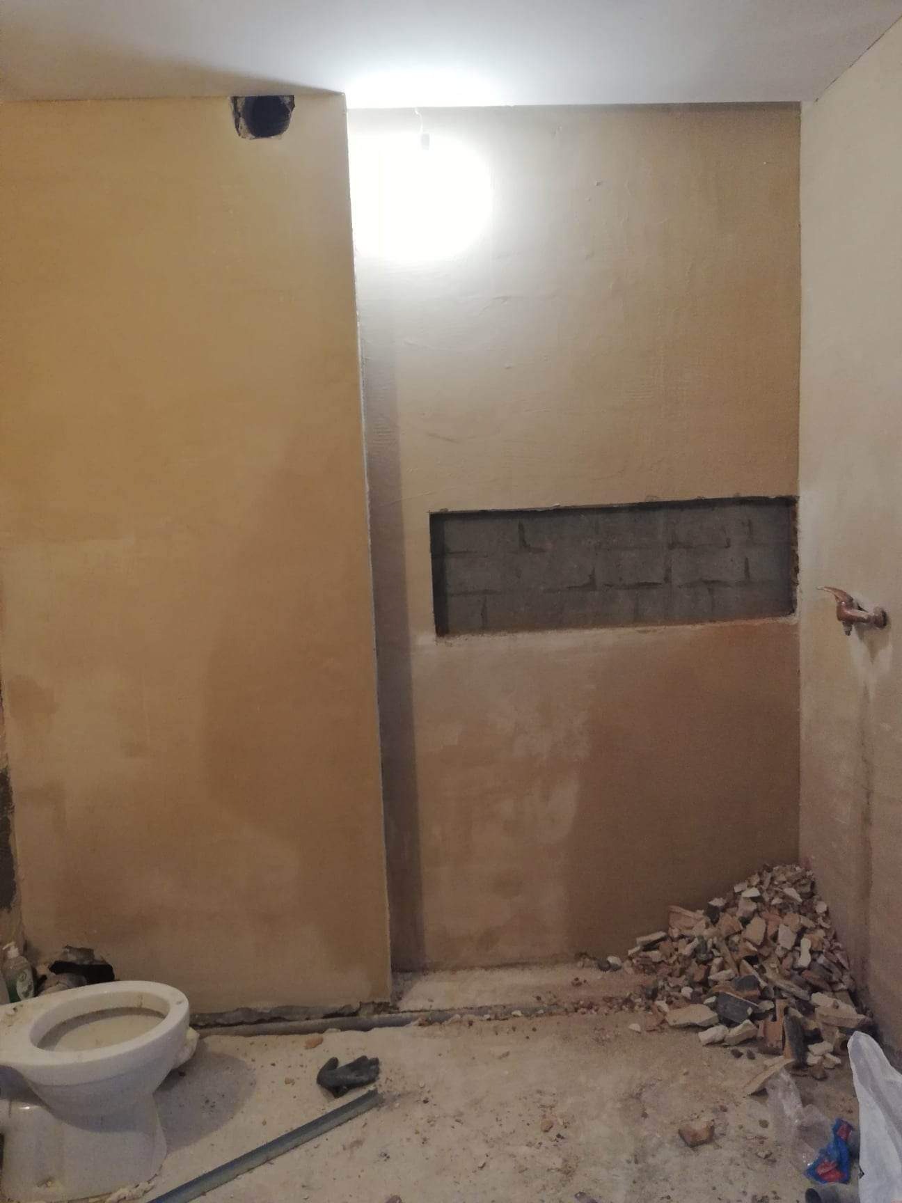 Łazienka w trakcie remontu: widoczne ściany z nierówną powierzchnią, otwór w ścianie z widocznymi cegłami, gruz na podłodze oraz częściowo zamontowana toaleta.