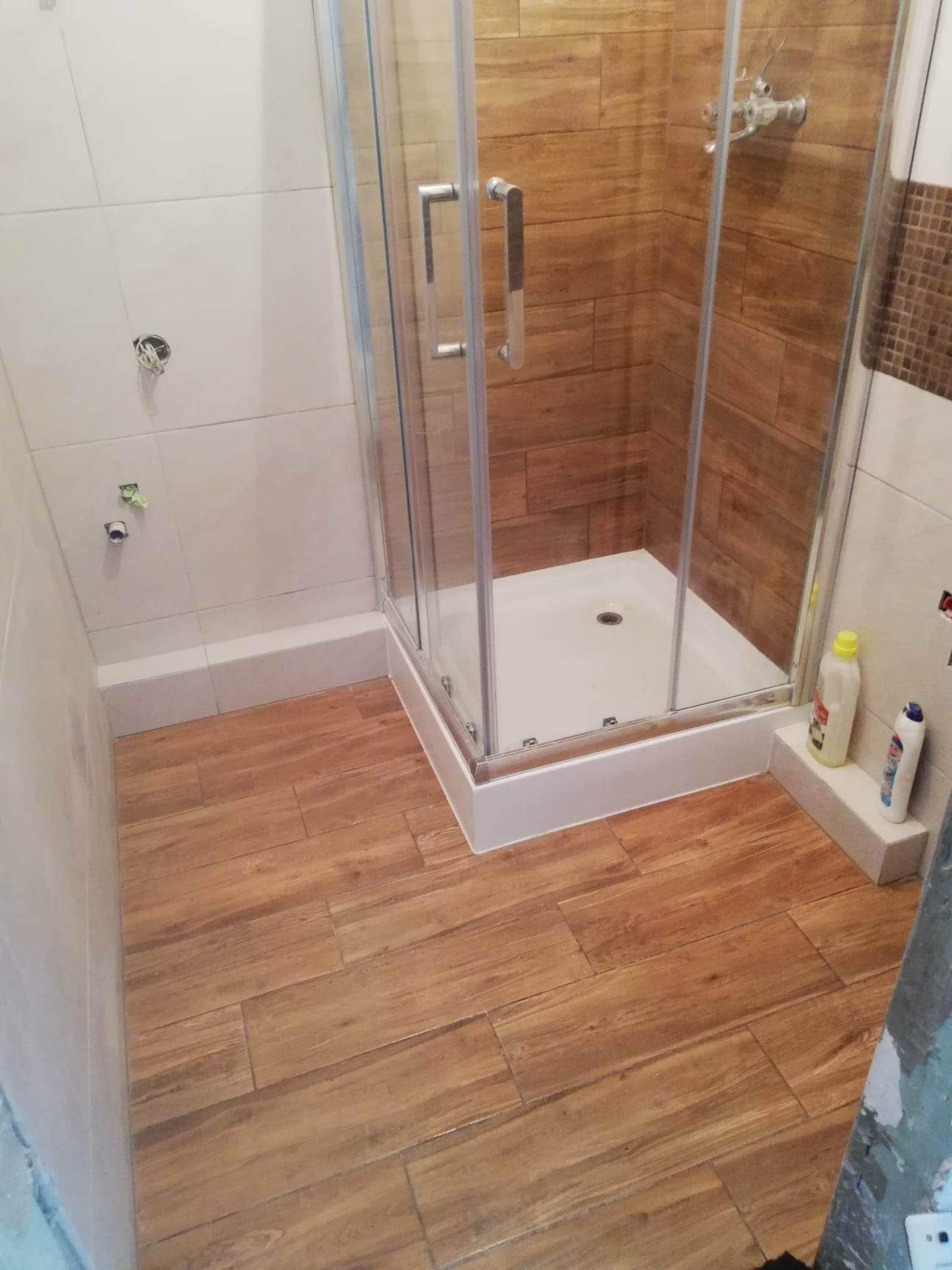 Wnętrze łazienki w trakcie prac wykończeniowych: kabina prysznicowa z chromowanymi uchwytami, płytki imitujące drewno na podłodze, częściowo wykończone ściany z białymi płytkami i wystającymi...