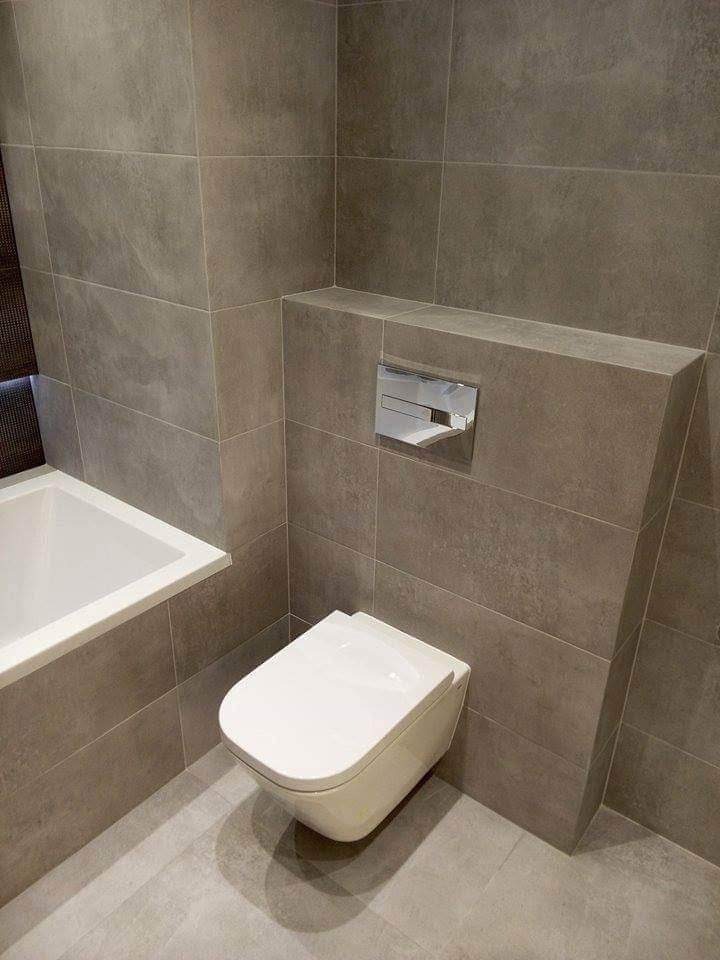 Nowoczesna łazienka z szarą płytką, wiszącą toaletą i częściowo zabudowanym systemem spłukiwania, widoczny fragment wanny.