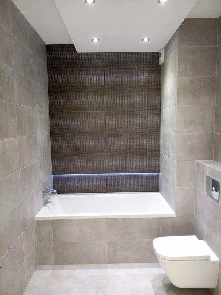 Nowoczesna łazienka z wanną obudowaną szarymi płytkami, podświetleniem LED nad wanną i wiszącą toaletą.