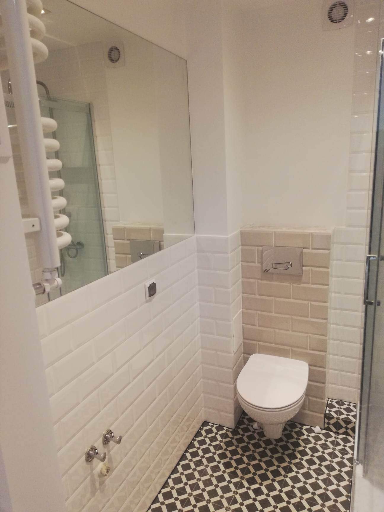 Wykończona łazienka z biało-beżowymi płytkami typu metro na ścianach, czarno-białą podłogą w geometryczny wzór, wiszącą toaletą i grzejnikiem drabinkowym widocznym w lustrze.