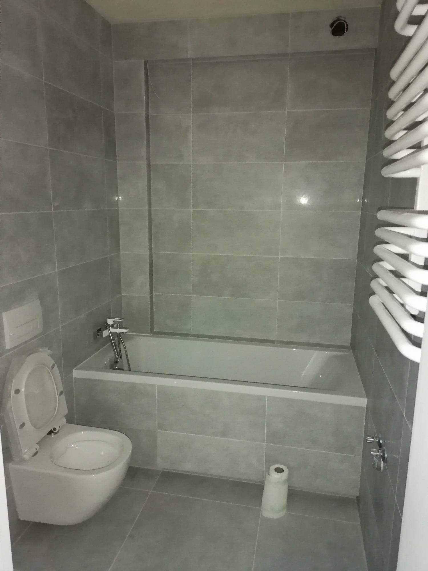 Nowoczesna łazienka wykończona szarymi płytkami, z wiszącą toaletą, wanną z baterią i grzejnikiem drabinkowym.