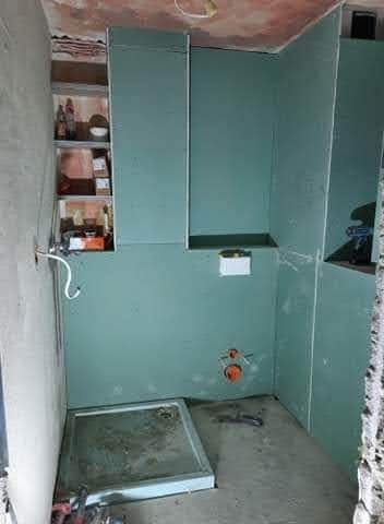 Wnętrze łazienki w trakcie prac: ściany wyłożone płytami gipsowo-kartonowymi, widoczna wnęka z półkami, przygotowana instalacja wodno-kanalizacyjna i brodzik.