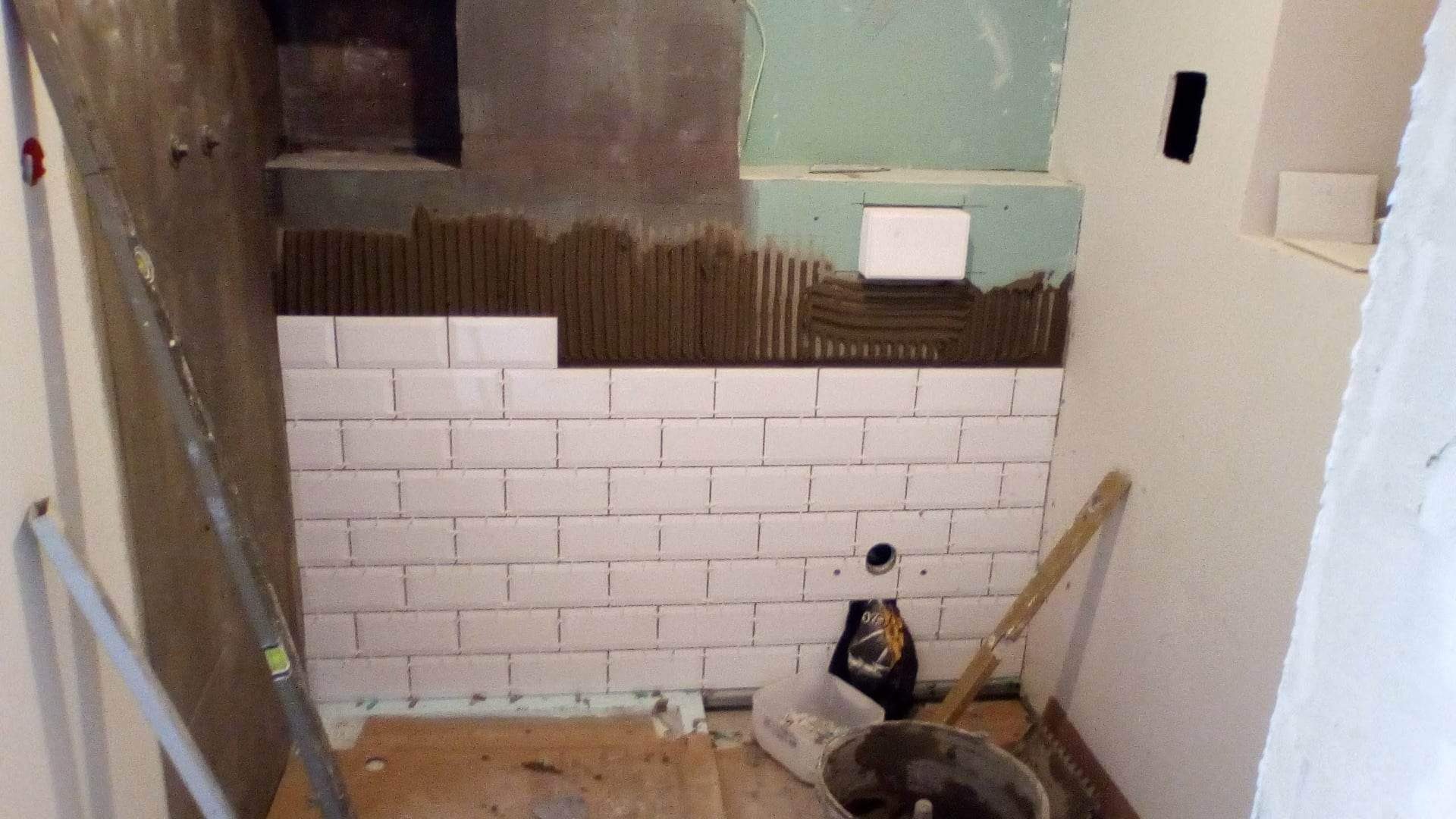 Białe, prostokątne płytki ułożone na ścianie łazienki, z widocznym klejem pod kolejnymi rzędami, poziomica oparta o ścianę, wiadro i inne narzędzia na podłodze.