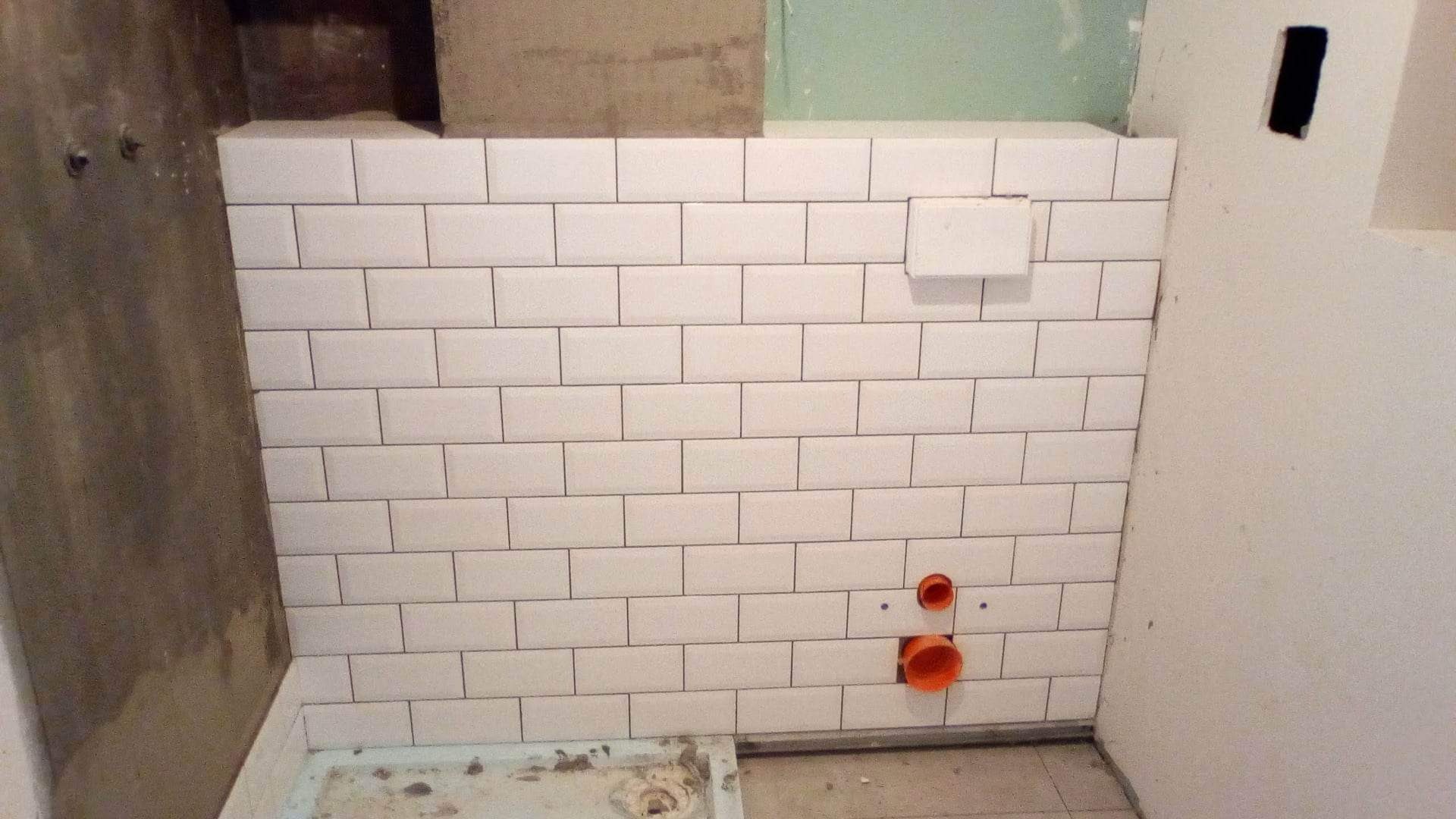 Białe, prostokątne płytki ułożone na ścianie łazienki, widoczne rury kanalizacyjne i fragment ściany w stanie surowym.