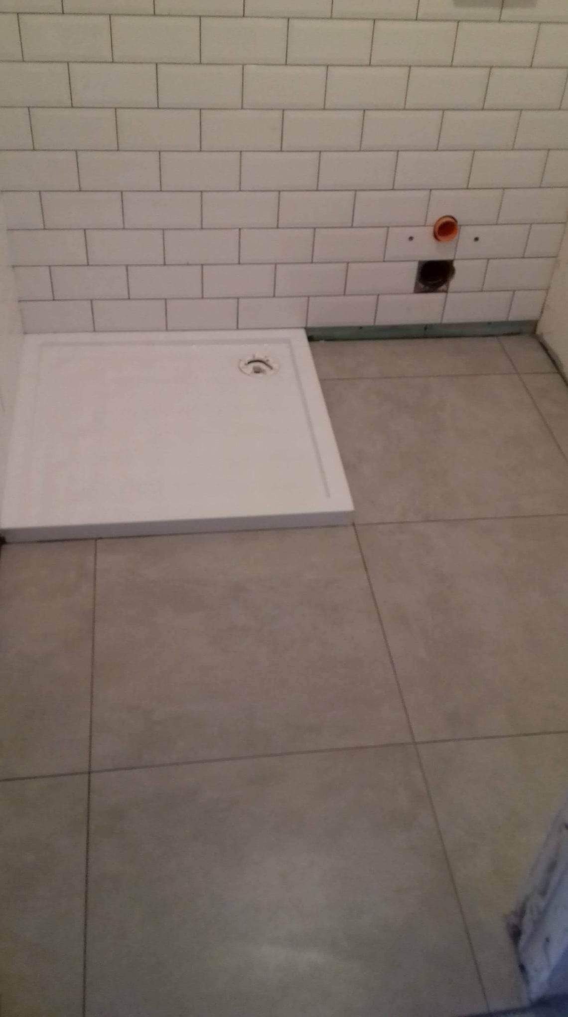 Biały brodzik posadzkowy w rogu łazienki, z widoczną instalacją hydrauliczną w ścianie z białych płytek, ułożone płytki podłogowe w odcieniach szarości.