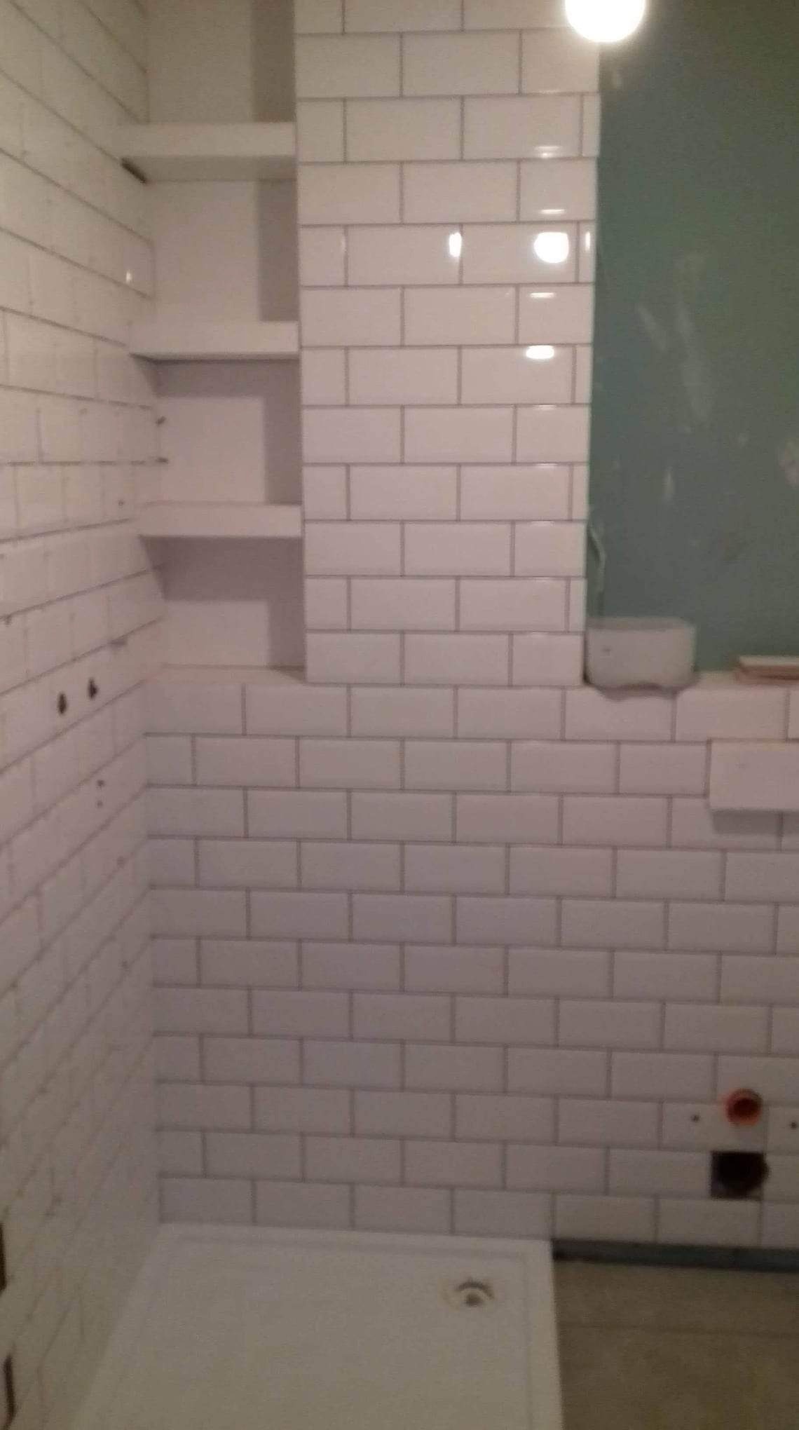 Ściana łazienki wyłożona białymi, prostokątnymi płytkami, z wnęką z półkami i widocznym brodzikiem prysznicowym. Widoczne niedociągnięcia w fugowaniu niektórych płytek.