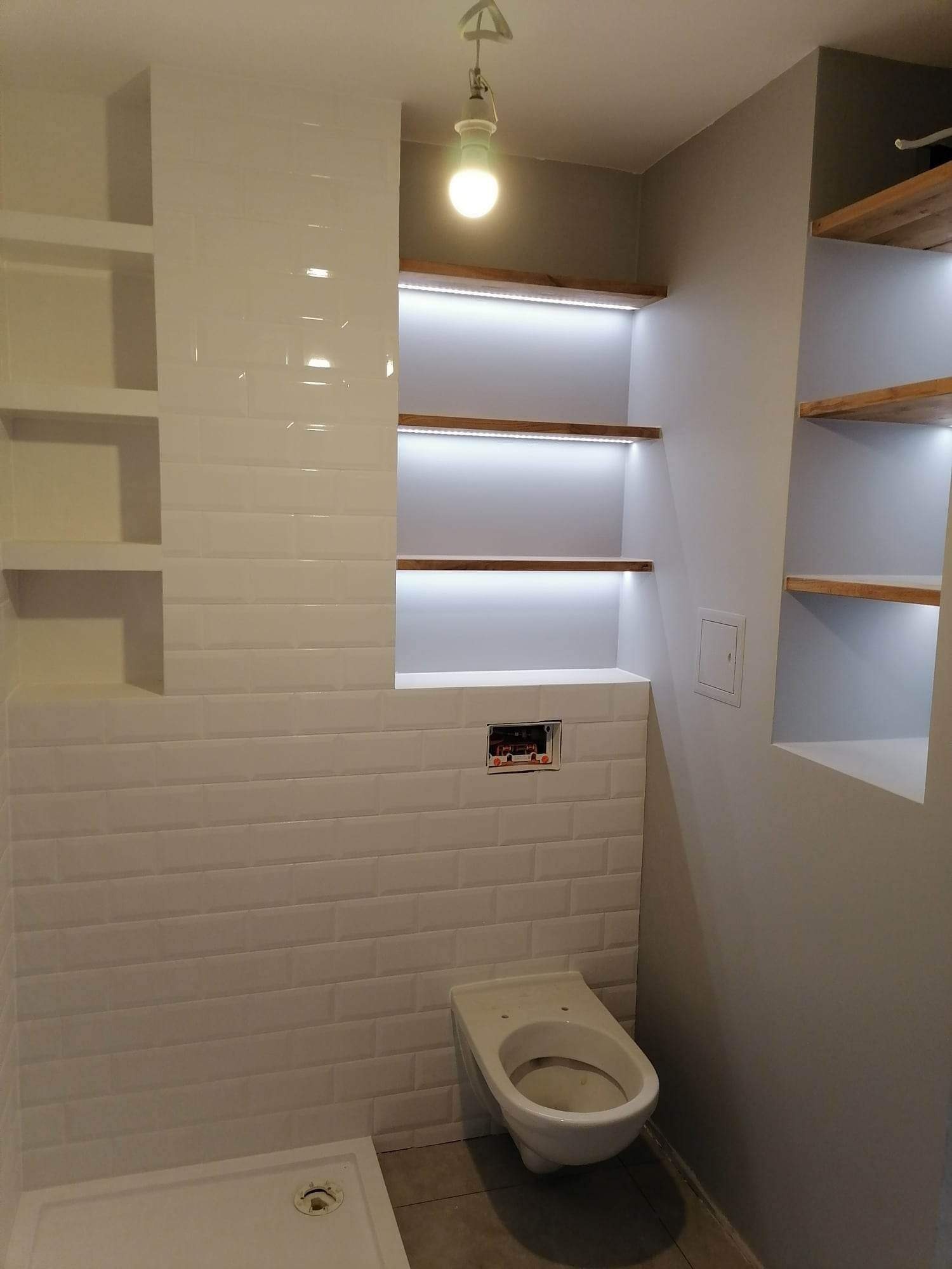 Wykończona łazienka z białymi, podłużnymi płytkami na ścianach, wbudowanymi półkami z oświetleniem LED i wiszącą toaletą.