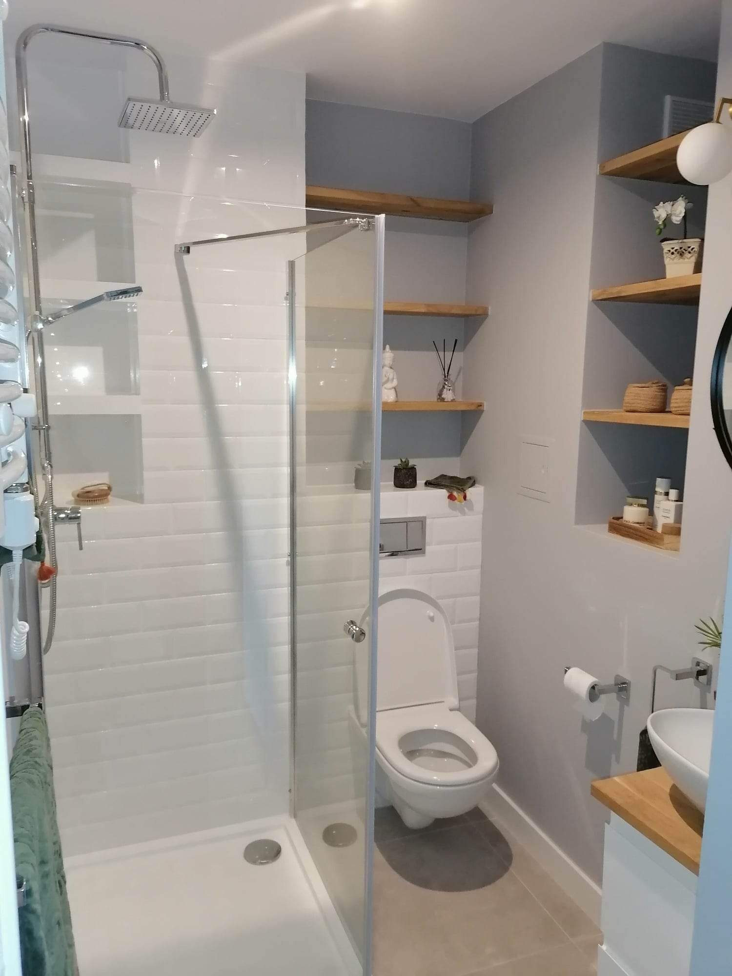 Nowoczesna łazienka z kabiną prysznicową typu walk-in, białą ceramiką, drewnianymi półkami i szarą ścianą, wykończona w minimalistycznym stylu.