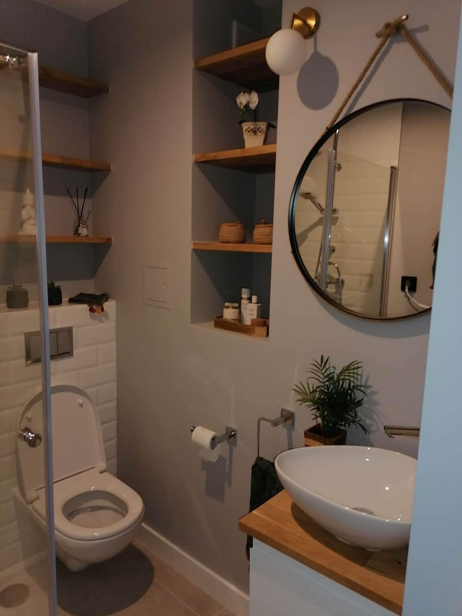 Nowoczesna łazienka w szarościach z elementami drewna: okrągłe lustro z liną, drewniane półki z dekoracjami, biała umywalka nablatowa i toaleta w kabinie prysznicowej.