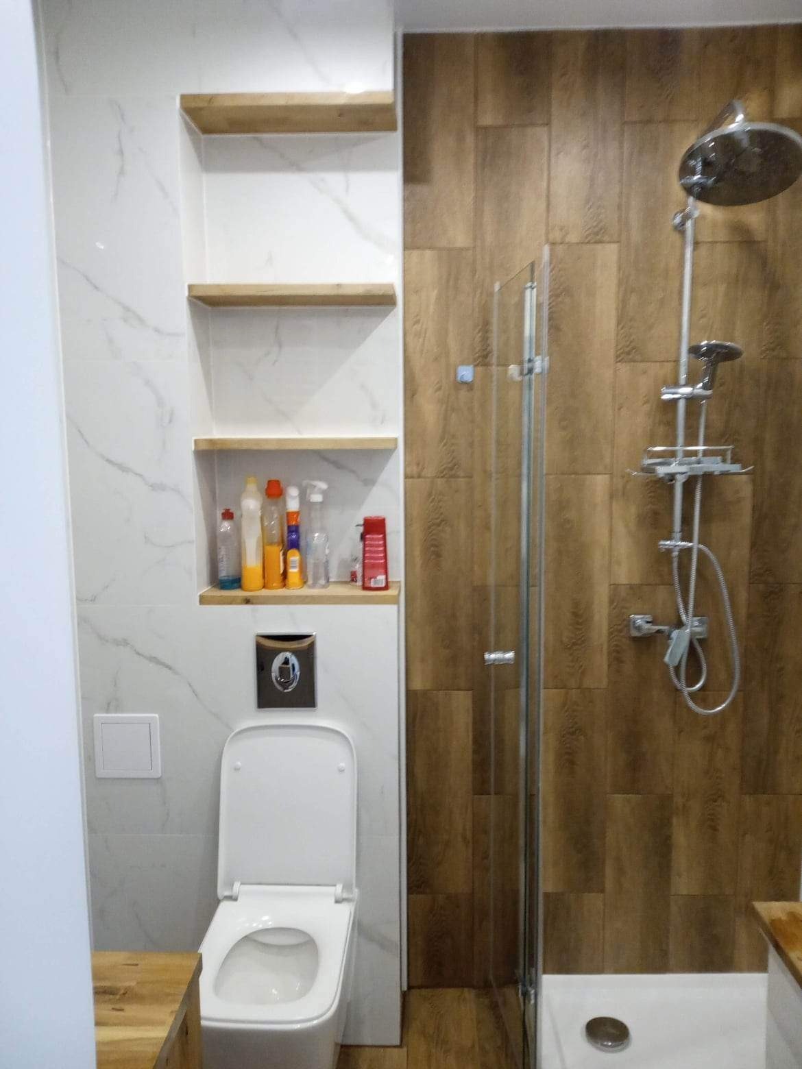 Wykończona łazienka z drewnopodobnymi płytkami w kabinie prysznicowej, toaleta z przyciskiem w kolorze grafitowym i wnęka z drewnianymi półkami zawierająca środki czystości.