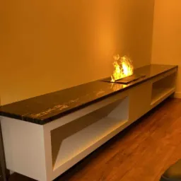 Długi, niski mebel z blatem z ciemnego kamienia z wbudowanym biokominkiem z żywym ogniem, na tle pomarańczowej ściany i drewnianej podłogi.