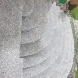 Granitowe schody zewnętrzne z zaokrąglonymi stopniami, zraszane wodą z konewki przez osobę w szortach, widok z g&oacute;ry.