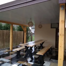 Zadaszony taras z drewnianym stołem i ławkami na posadzce z płytek kamiennych o różnych kolorach i fakturach, oświetlony wiszącymi lampami.