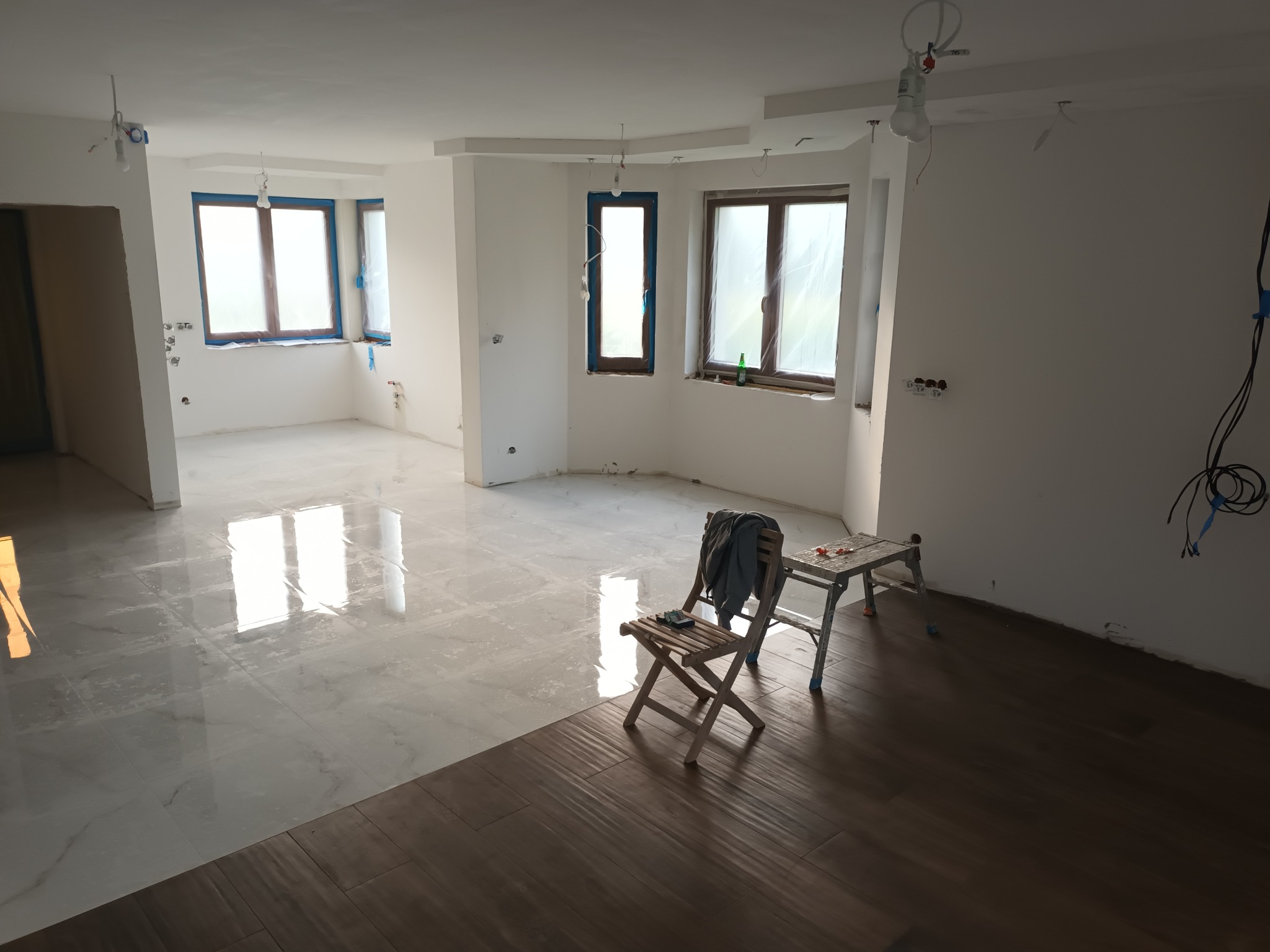 Wnętrze w trakcie prac wykończeniowych: część podłogi z jasnych, marmurkowych płytek, część z ciemnych paneli imitujących drewno; okna zabezpieczone folią, widoczne narzędzia i drabina.