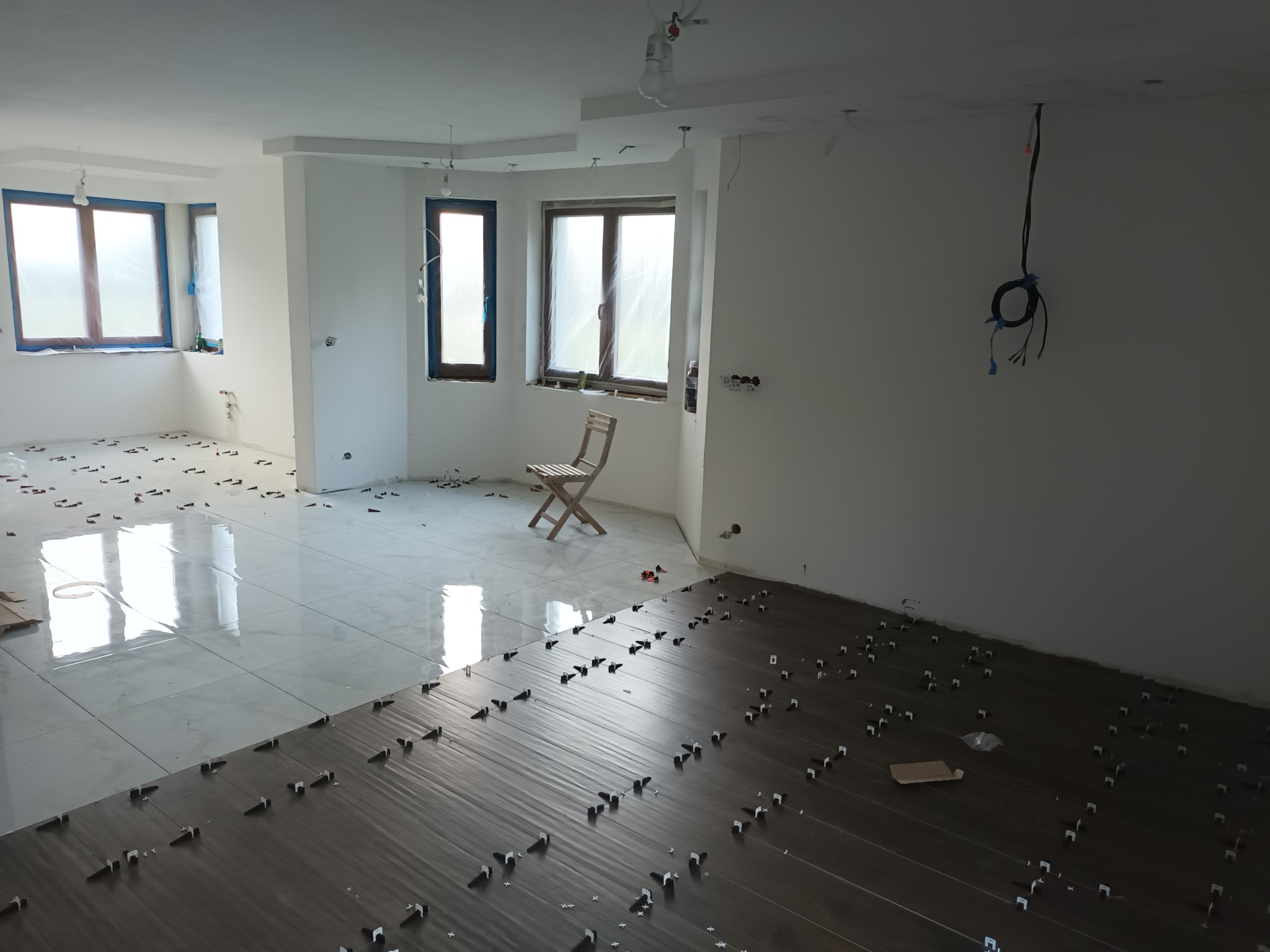 Wnętrze w trakcie prac wykończeniowych: częściowo ułożone jasne płytki podłogowe, fragment podkładu pod ciemne panele z widocznymi elementami poziomującymi, okna zabezpieczone folią, surowe ściany,...