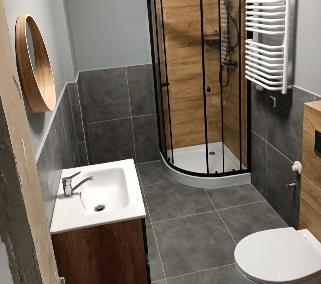 Szara łazienka z kabiną prysznicową z czarnymi profilami, okrągłym lustrem z drewnianą ramą i umywalką z szafką w odcieniu drewna, płytki podłogowe i ścienne w odcieniach szarości.