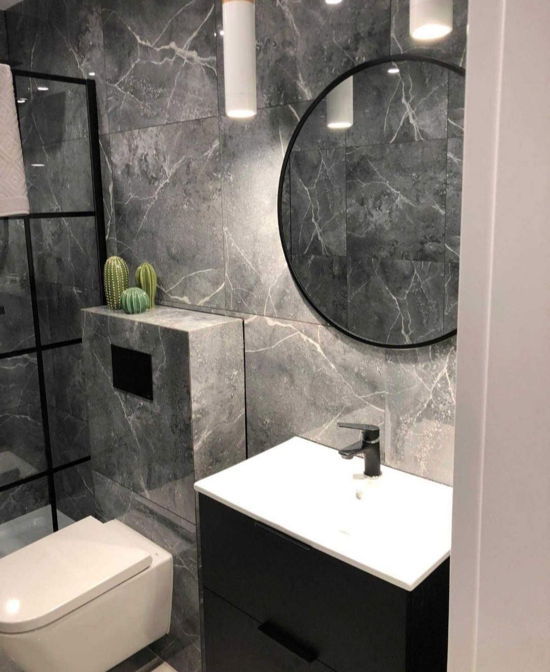 Nowoczesna łazienka z szarymi płytkami imitującymi marmur, okrągłym lustrem w czarnej ramie, podwieszaną toaletą, czarną szafką pod umywalką i dekoracyjnymi kaktusami na zabudowie stelaża.