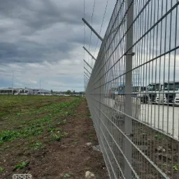 Długa, metalowa siatka ogrodzeniowa z drutem kolczastym na szczycie, oddzielająca teren zielony od placu z zaparkowanymi ciężarówkami, sfotografowana pod pochmurnym niebem.