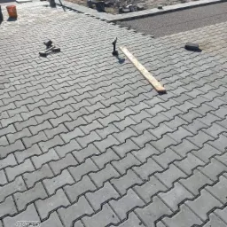 Fragment układanej szarej kostki brukowej typu 'I', widoczne narzędzia brukarskie: młotek, poziomica, oraz drewniana listwa, w tle przygotowane podłoże z kruszywa.