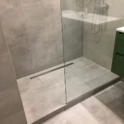 Nowoczesna kabina prysznicowa walk-in z odpływem liniowym i szklaną ścianką, wykończona szarymi płytkami wielkoformatowymi, z widoczną zieloną szafką łazienkową i fragmentem wanny w tle.