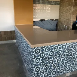 Bar w trakcie wykańczania, z blatem w kolorze beżowym i ścianami obłożonymi dekoracyjnymi, niebiesko-białymi płytkami w marokańskim stylu. W tle widoczna ściana z częściowo ułożonymi, brązowymi...