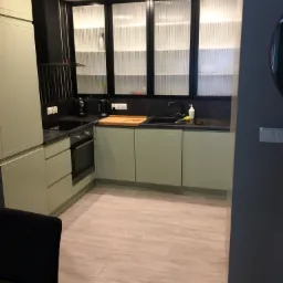 Nowoczesna kuchnia w odcieniach zieleni z frezowanymi frontami szafek, czarnym blatem i szklanymi szafkami górnymi z ryflowanym szkłem, oświetlona punktowo