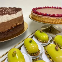 Elegancka kompozycja deserów: tort trójwarstwowy, tarta z malinami i mini deserki pistacjowe na złotej tacy.