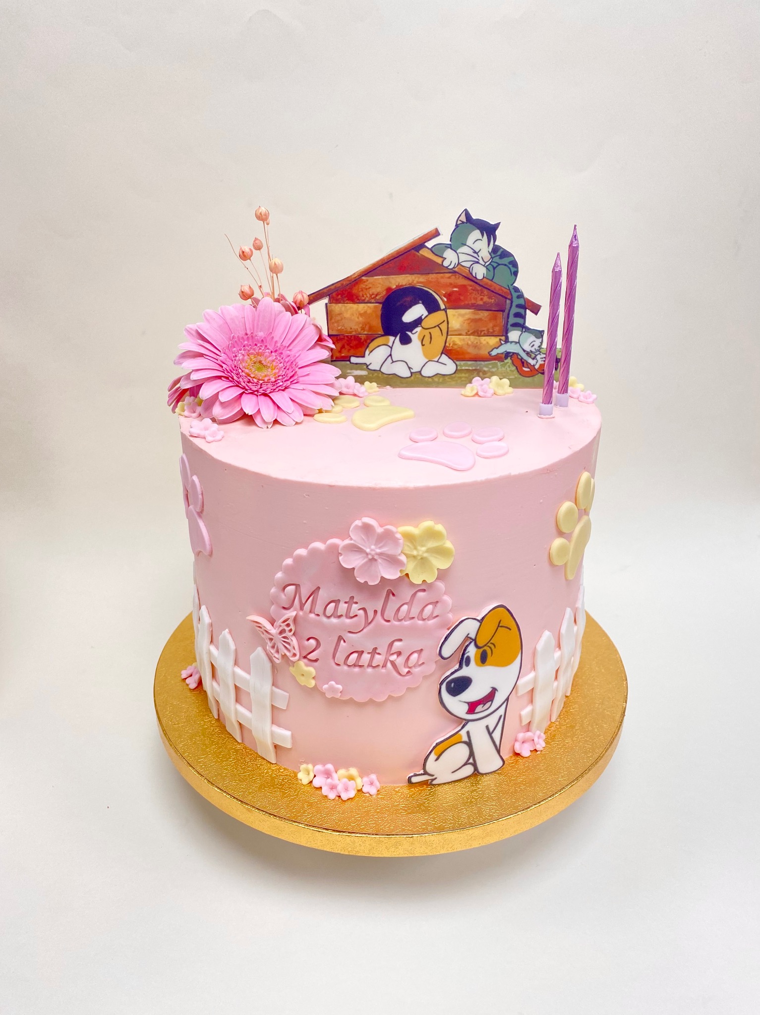 Różowy tort urodzinowy z motywem bajkowym: pies w budzie, koty, napis 'Matylda 2 latka', dekoracje z kwiatów i płotków na złotym podkładzie.