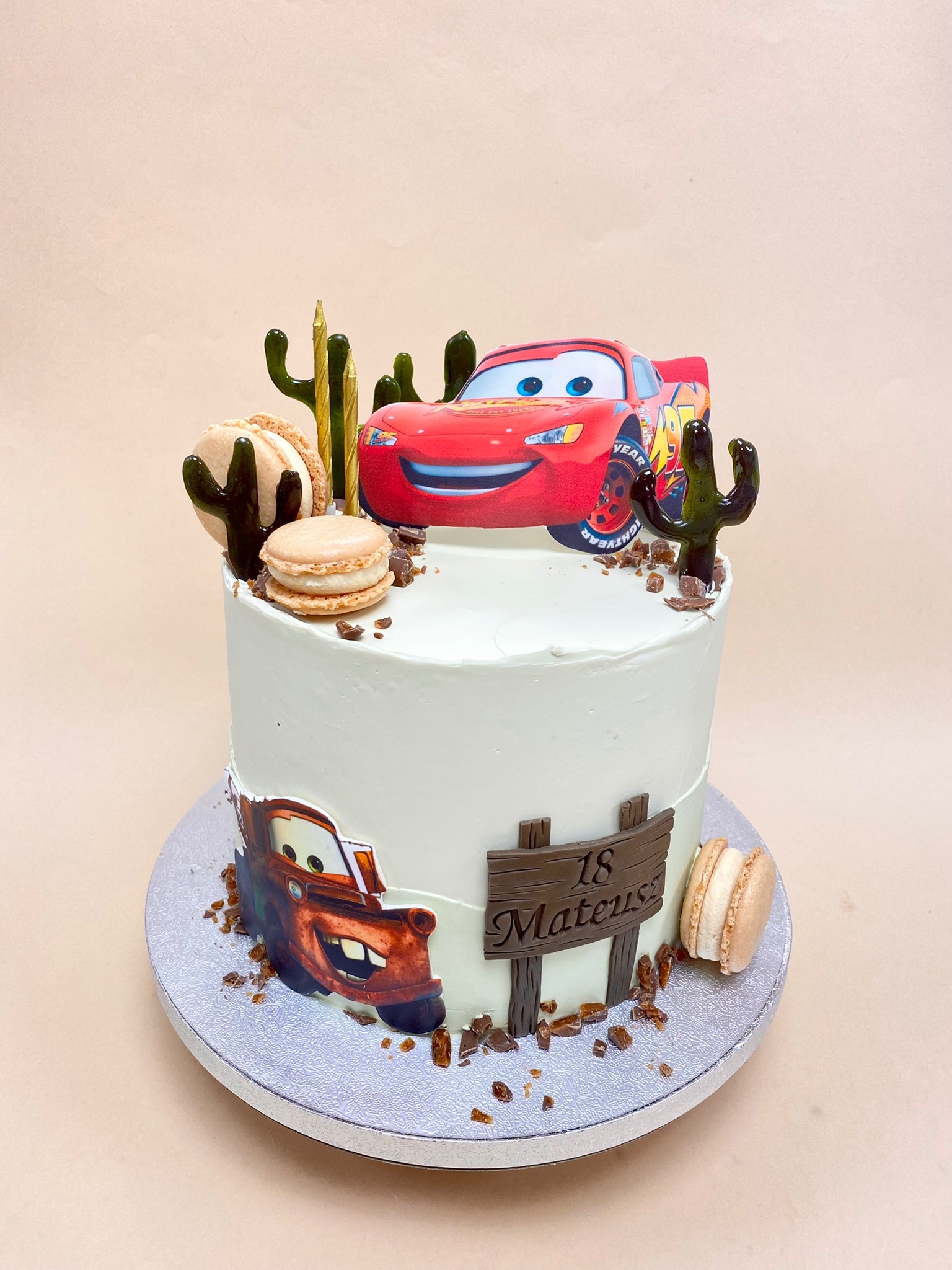 Tort urodzinowy z motywem Aut, dekorowany figurką Zygzaka McQueen, kaktusami z lukru, makaronikami i tabliczką z napisem '18 Mateusz'.