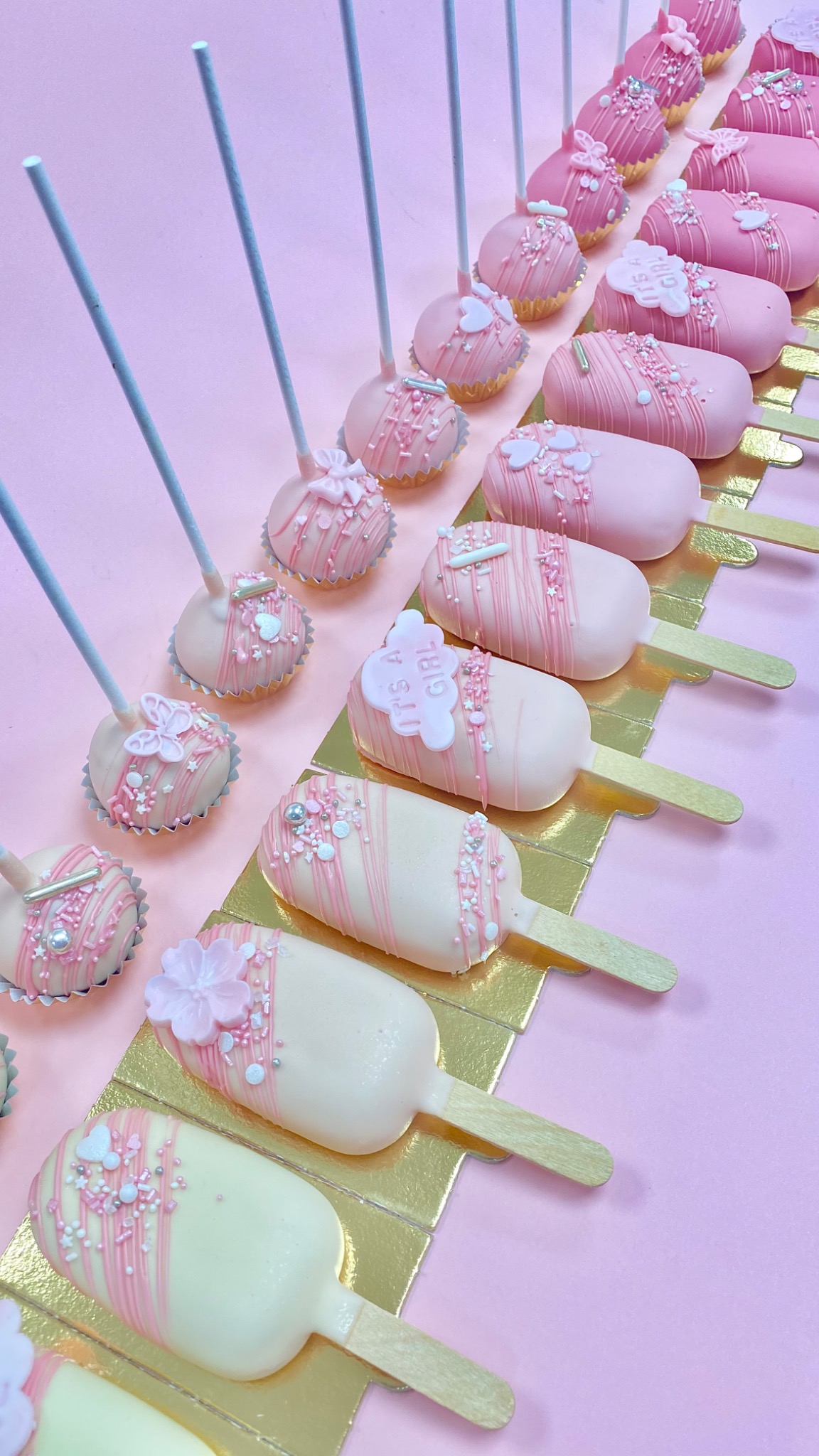 Rząd jasnoróżowych cake pops i cake sicles z jasnoróżowymi dekoracjami, w tym posypkami, kwiatami i napisem 'It's a Girl', ułożonych na złotej tacy na jasnoróżowym tle.