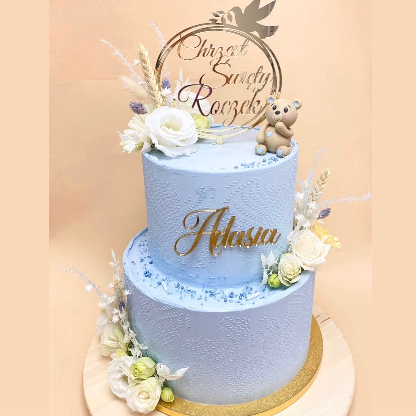 Dwupoziomowy tort w odcieniach błękitu z delikatnym tłoczeniem, zdobiony białymi kwiatami, suszonymi trawami, złotym napisem 'Adasia', topperem 'Chrzest Święty Roczek' i figurką misia.