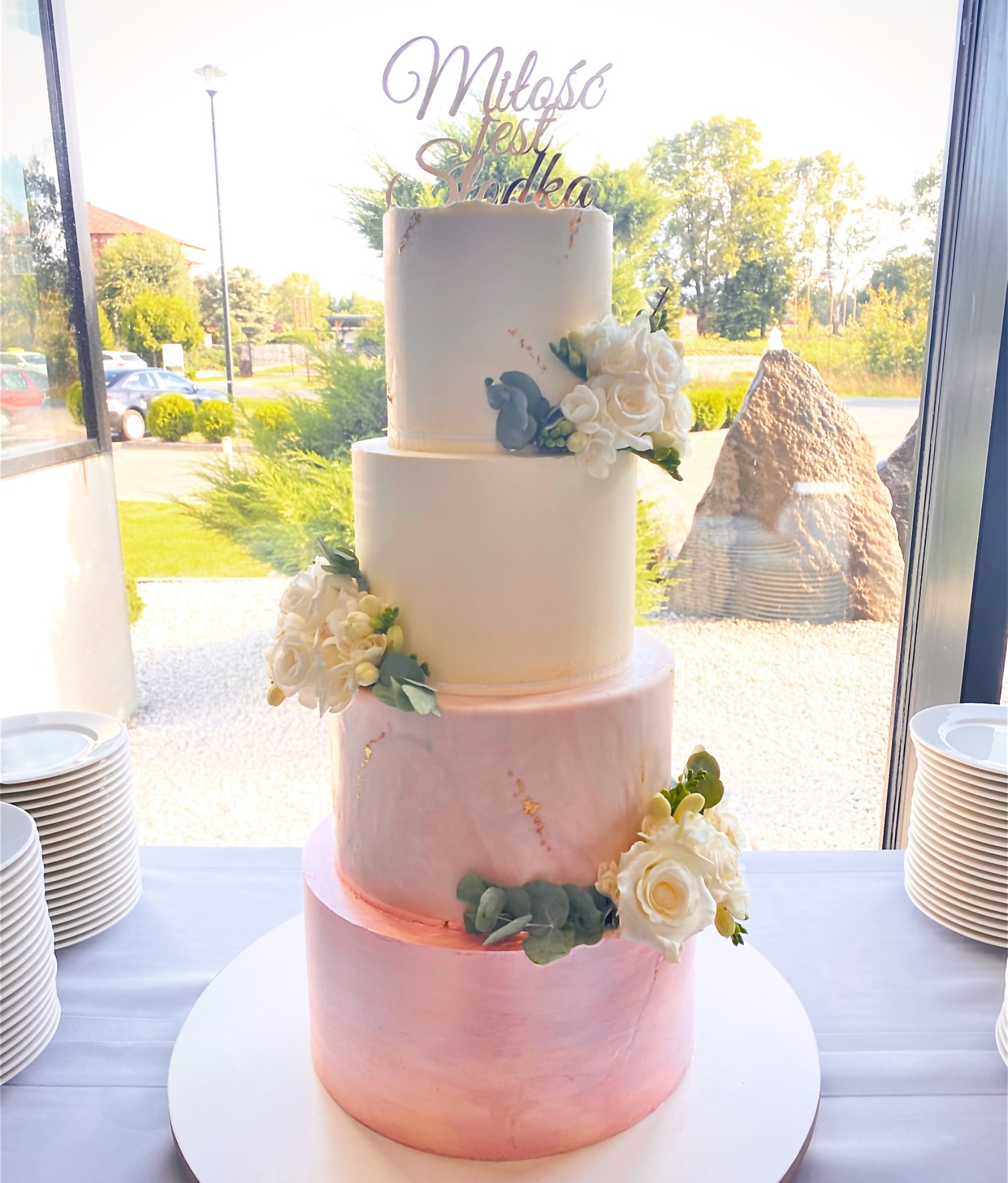 Elegancki tort weselny, czteropiętrowy, w odcieniach bieli i różu, zdobiony żywymi białymi kwiatami i złotymi akcentami, z napisem 'Miłość jest słodka' na szczycie, ustawiony na białym stole...