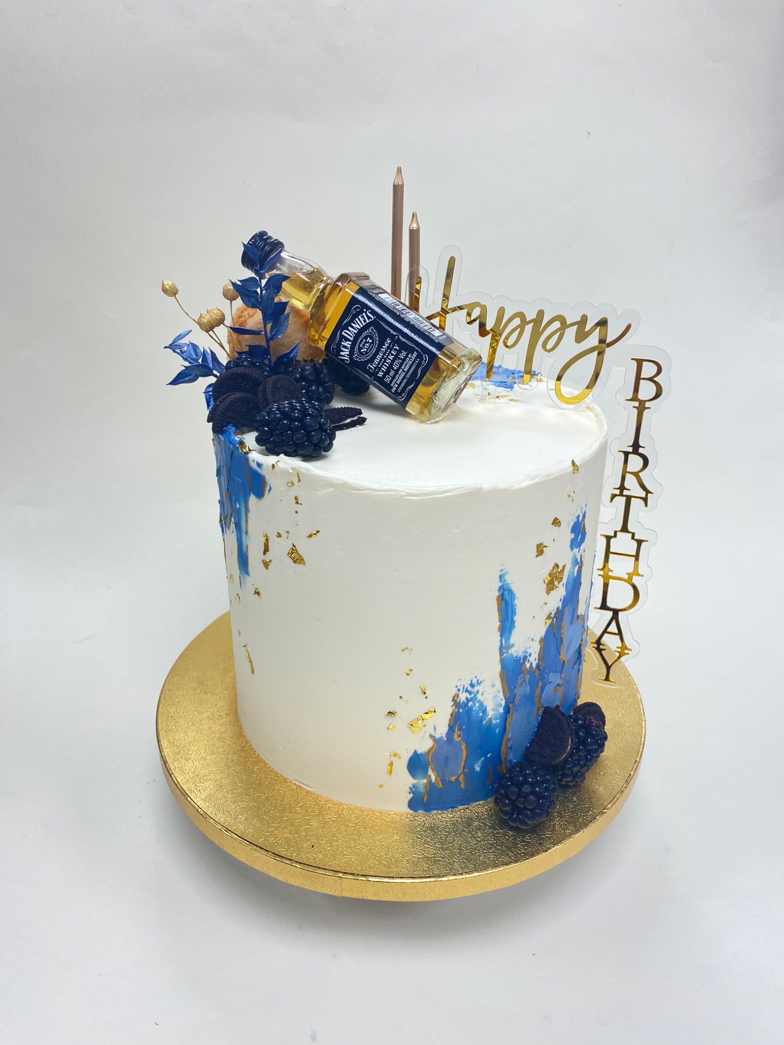 Tort urodzinowy z białym kremem, dekorowany niebieskimi akcentami, złotymi płatkami, jeżynami, ciastkami Oreo, butelką Jack Daniel's i napisem 'Happy Birthday'.