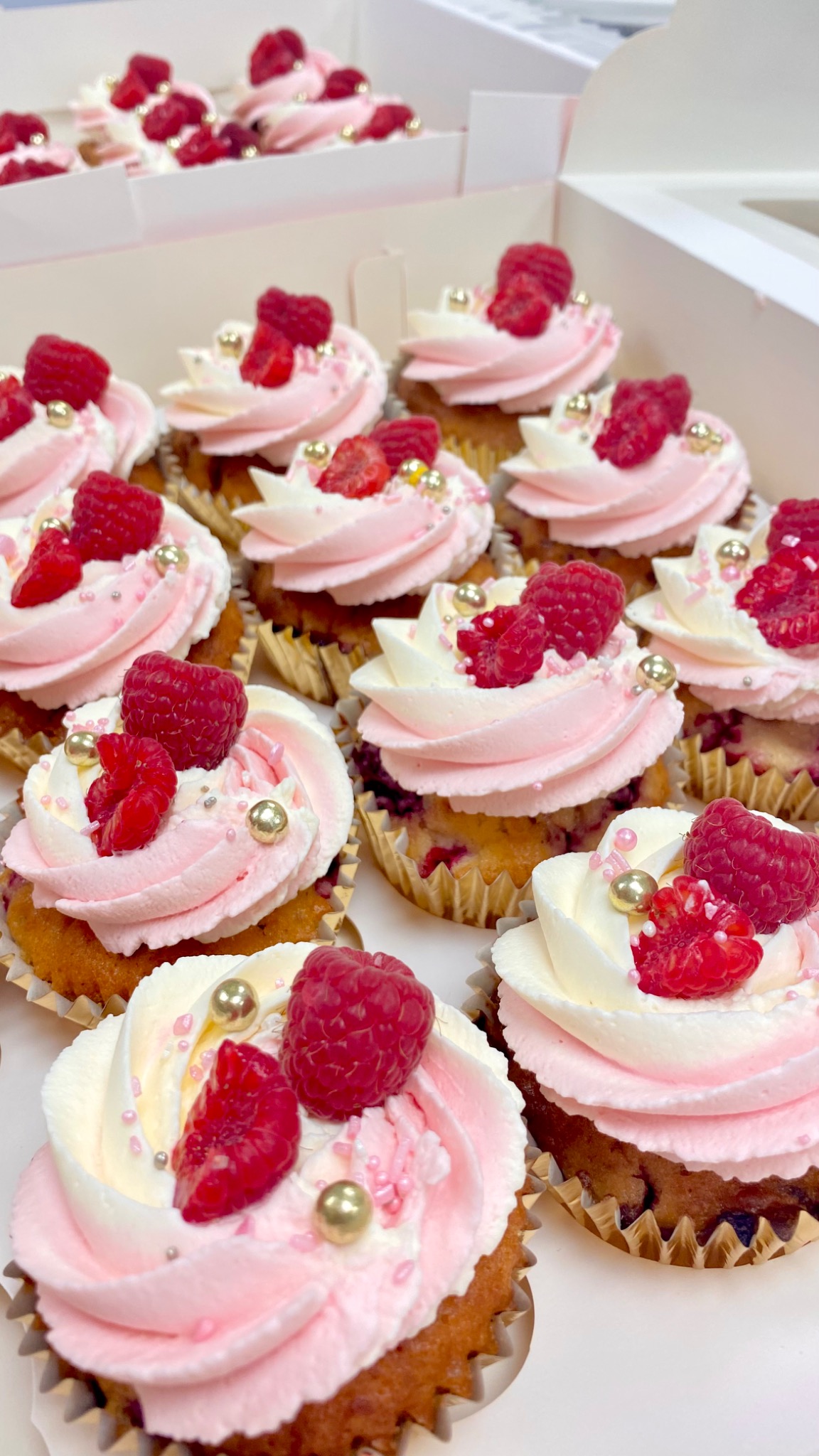 Rząd babeczek z różowym i białym kremem, malinami i złotymi kuleczkami, umieszczonych w białym pudełku.