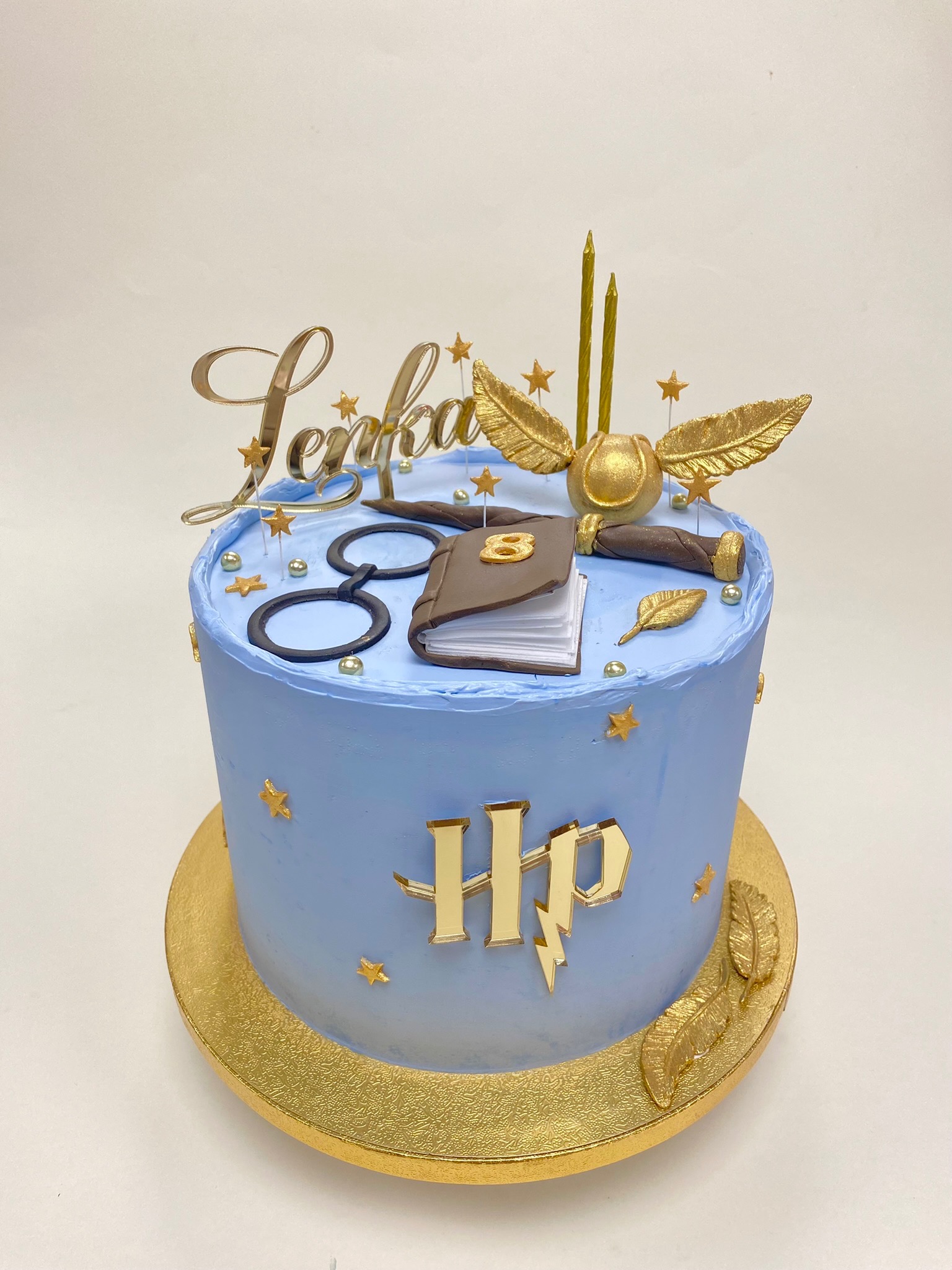 Tort urodzinowy w stylu Harry'ego Pottera z błękitnym kremem, złotymi dekoracjami: imieniem, insygniami, okularami, książką i Złotym Zniczem.
