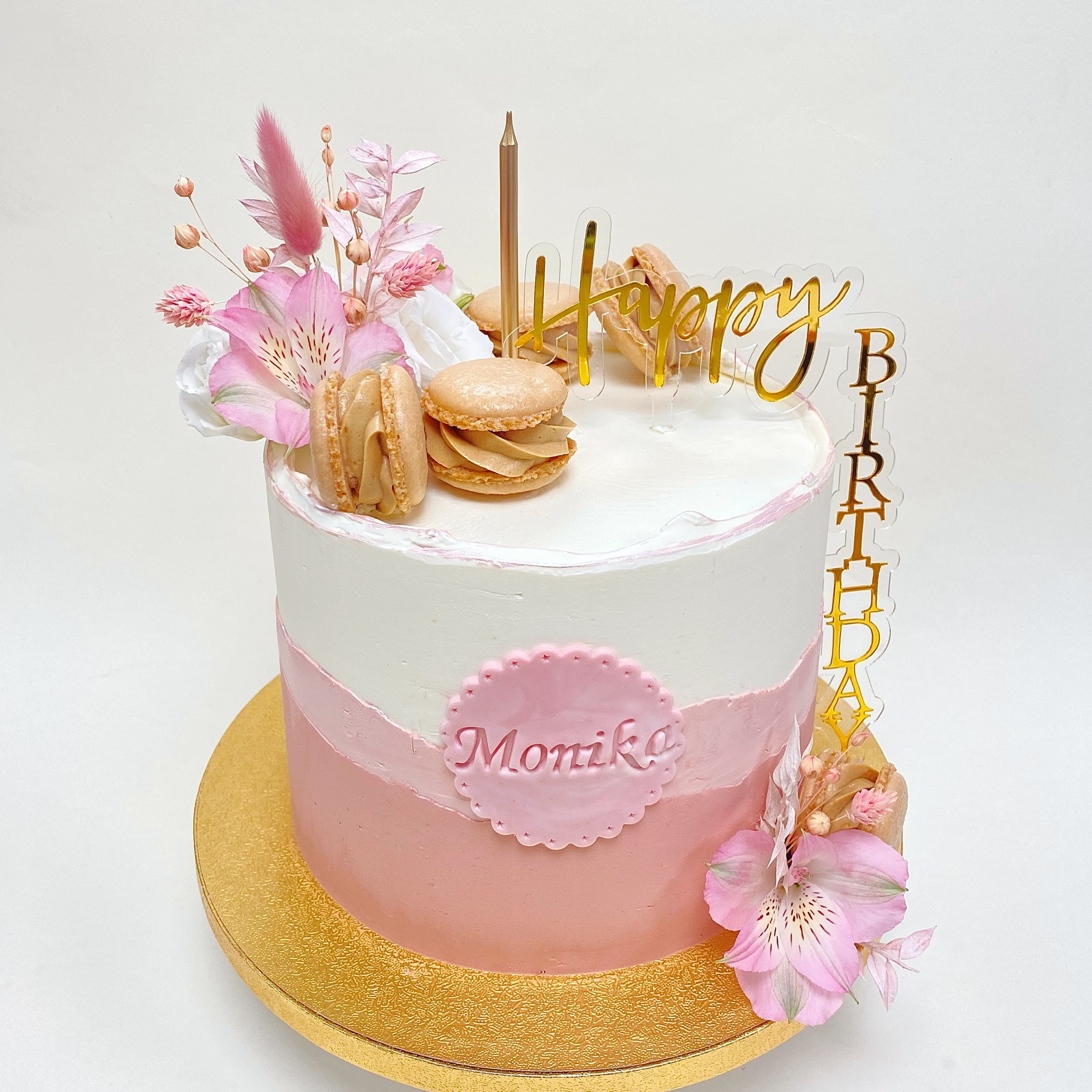 Tort dwukolorowy (biało-różowy) z napisem 'Monika', dekorowany kwiatami, makaronikami i złotym napisem 'Happy Birthday', umieszczony na złotej paterze.