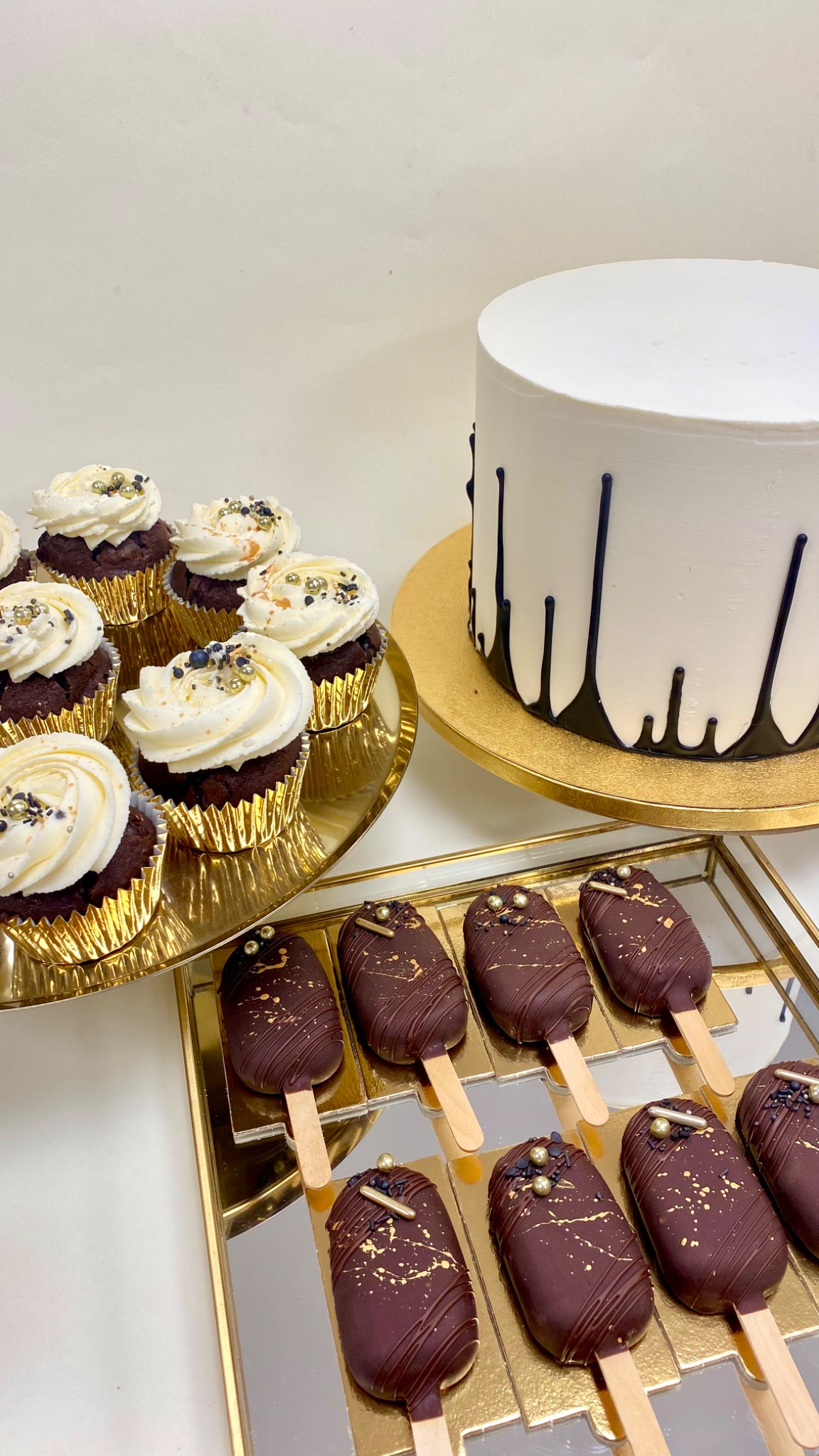 Elegancka kompozycja słodkości: czekoladowe babeczki z kremem, tort z czarnymi spływami i cake pops na patyku, wszystko na złotych podstawkach.