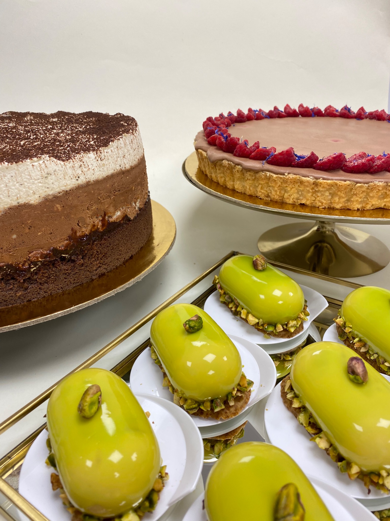 Elegancka kompozycja deserów: tort trójwarstwowy, tarta z malinami i mini deserki pistacjowe na złotej tacy.