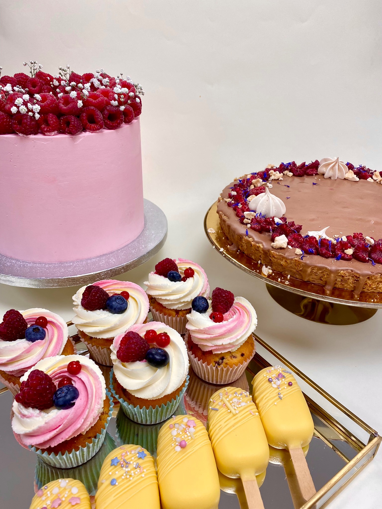 Różowy tort z malinami i gipsówką, czekoladowa tarta z bezami i kwiatami, babeczki z kremem i owocami oraz żółte cake pops na złotej tacy.