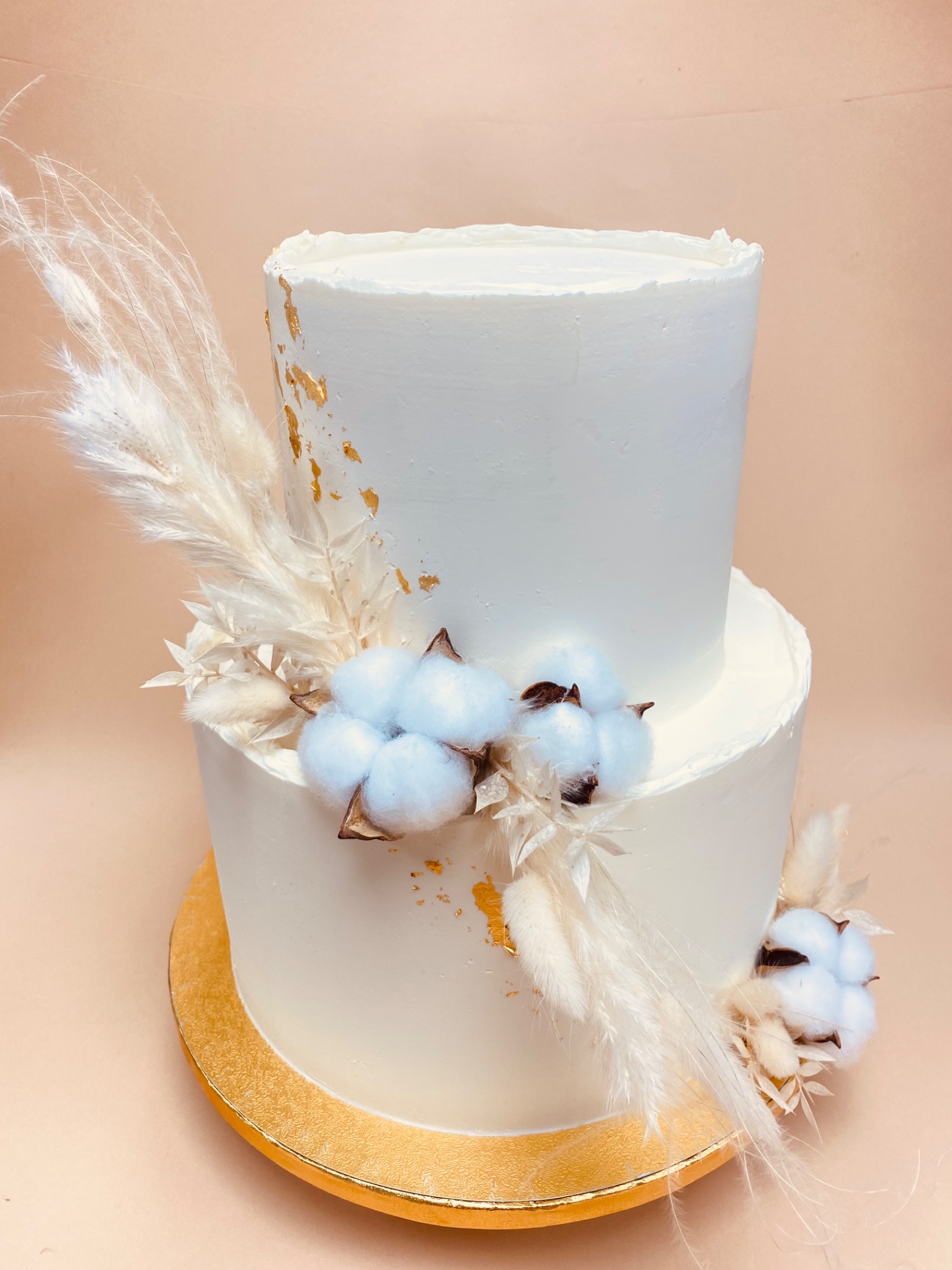 Dwupoziomowy tort weselny w białym kremie, zdobiony suszonymi kwiatami bawełny i złotymi płatkami, umieszczony na złotej podstawie.
