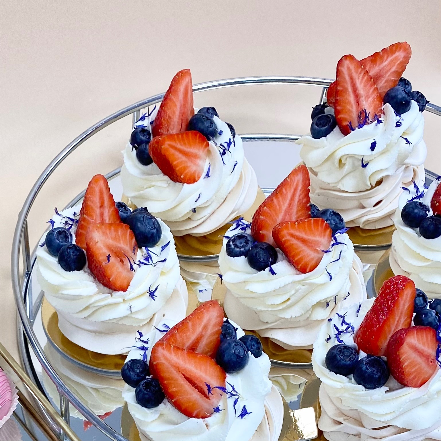 Mini bezy Pavlova udekorowane bitą śmietaną, świeżymi truskawkami, borówkami i drobnymi niebieskimi kwiatkami, ułożone na srebrnej paterze.