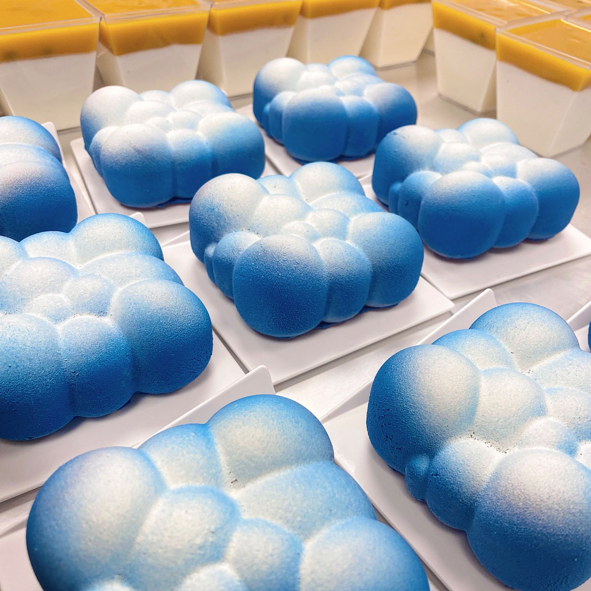 Niebiesko-białe desery w kształcie chmur na białych talerzykach, w tle deser w przezroczystych kubeczkach z żółtą warstwą na górze.