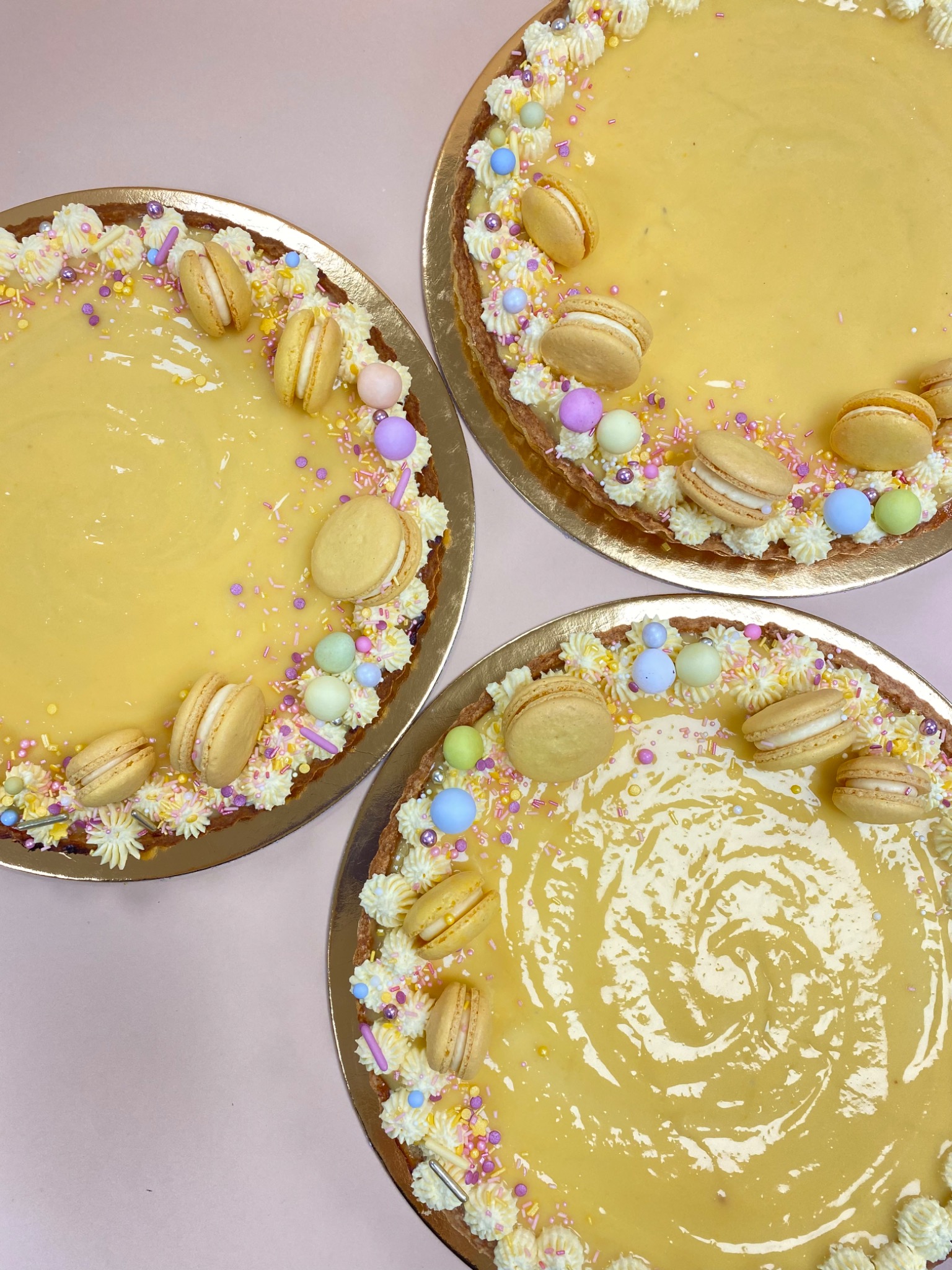 Trzy cytrynowe tarty na złotych podkładkach, udekorowane makaronikami, kremem i kolorowymi posypkami, ułożone na różowym tle.