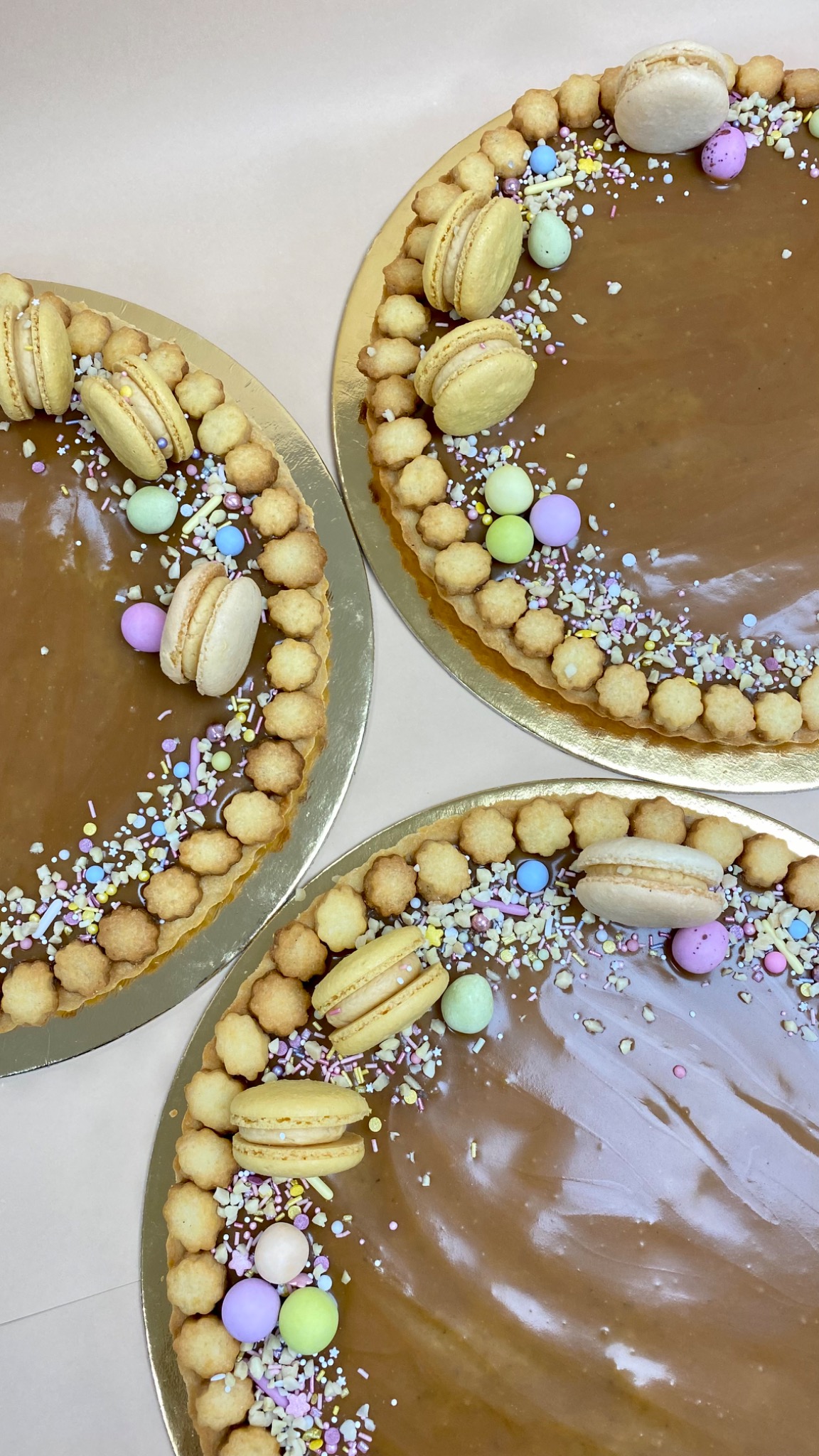 Trzy okrągłe tarty z karmelem, dekorowane mini ciasteczkami, makaronikami i kolorowymi kuleczkami, oferowane przez cukiernię w Sokolniki-Las.